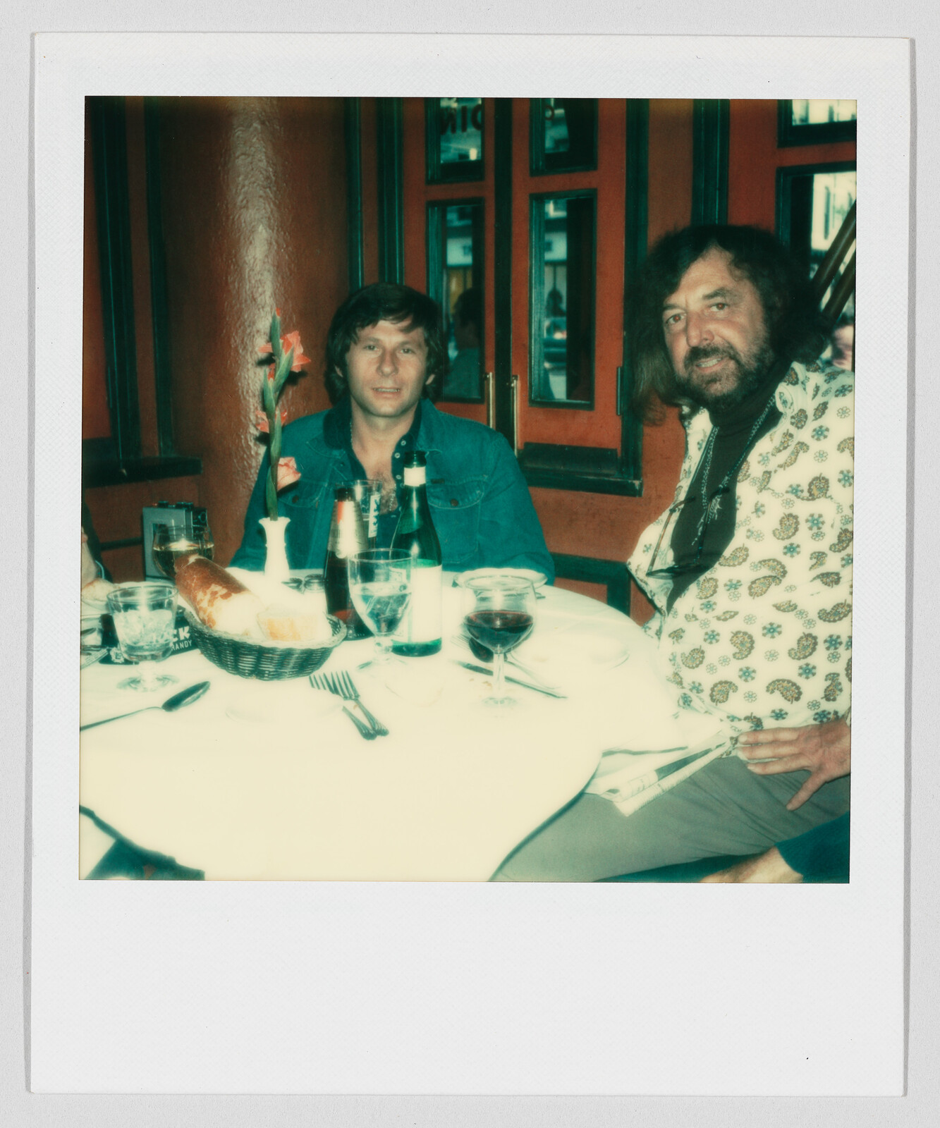 Two men sit at a restaurant table with wine, bottles, bread, and a single tall flower.