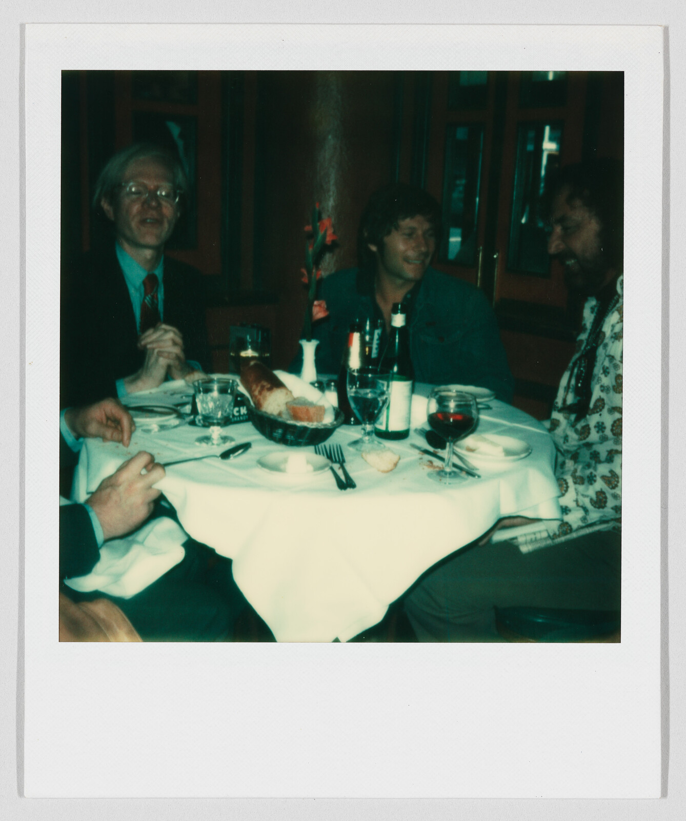 Three people sit around a restaurant table laughing and sharing bread, wine, and drinks.