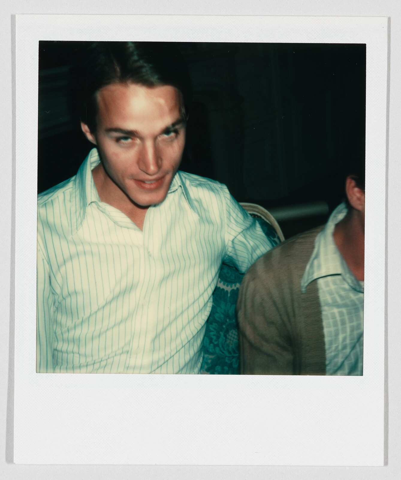 A young man in a light striped shirt sits on a patterned chair and looks toward the camera.