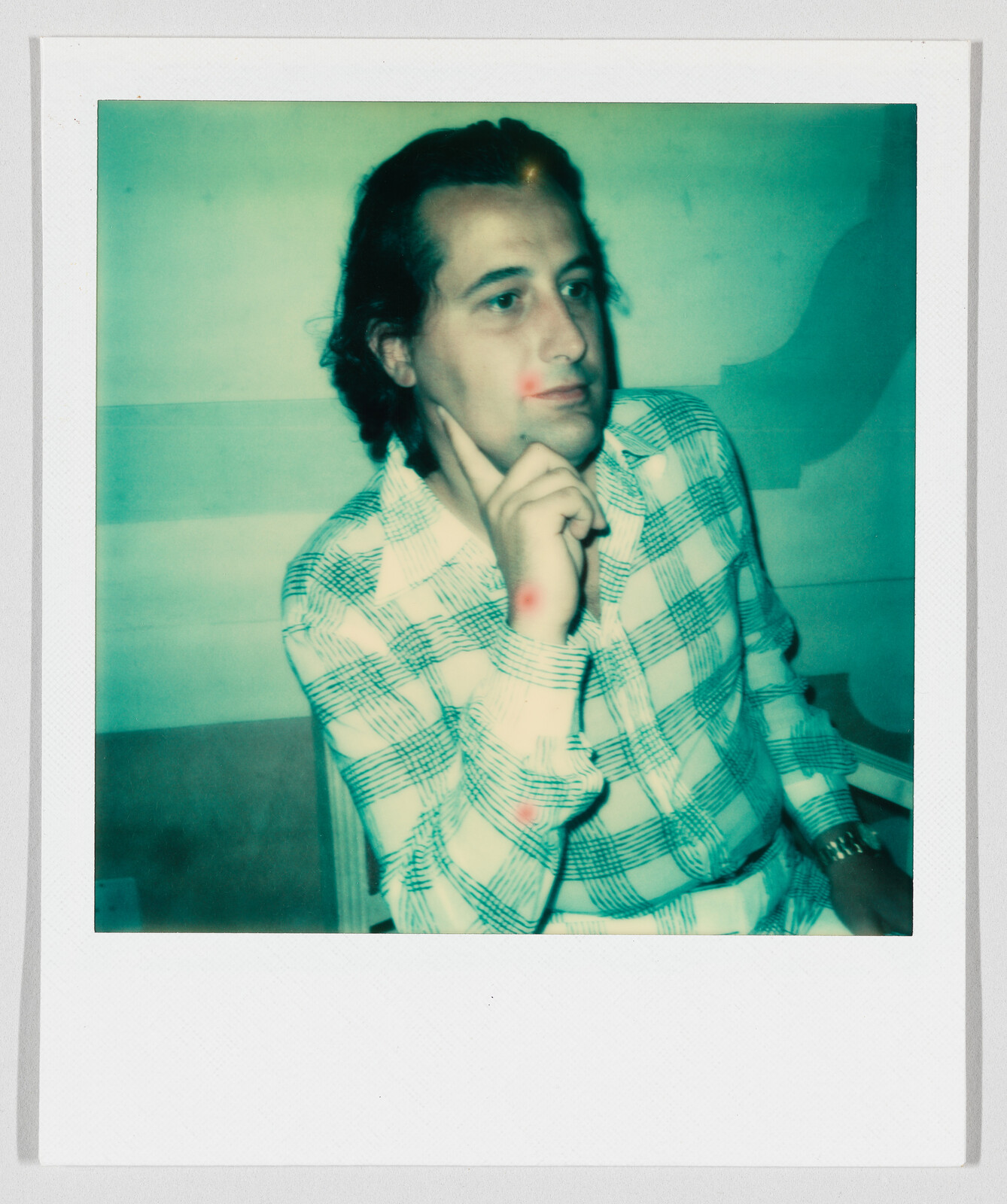 A man wearing a plaid shirt sits with his hand on his chin, looking thoughtful.