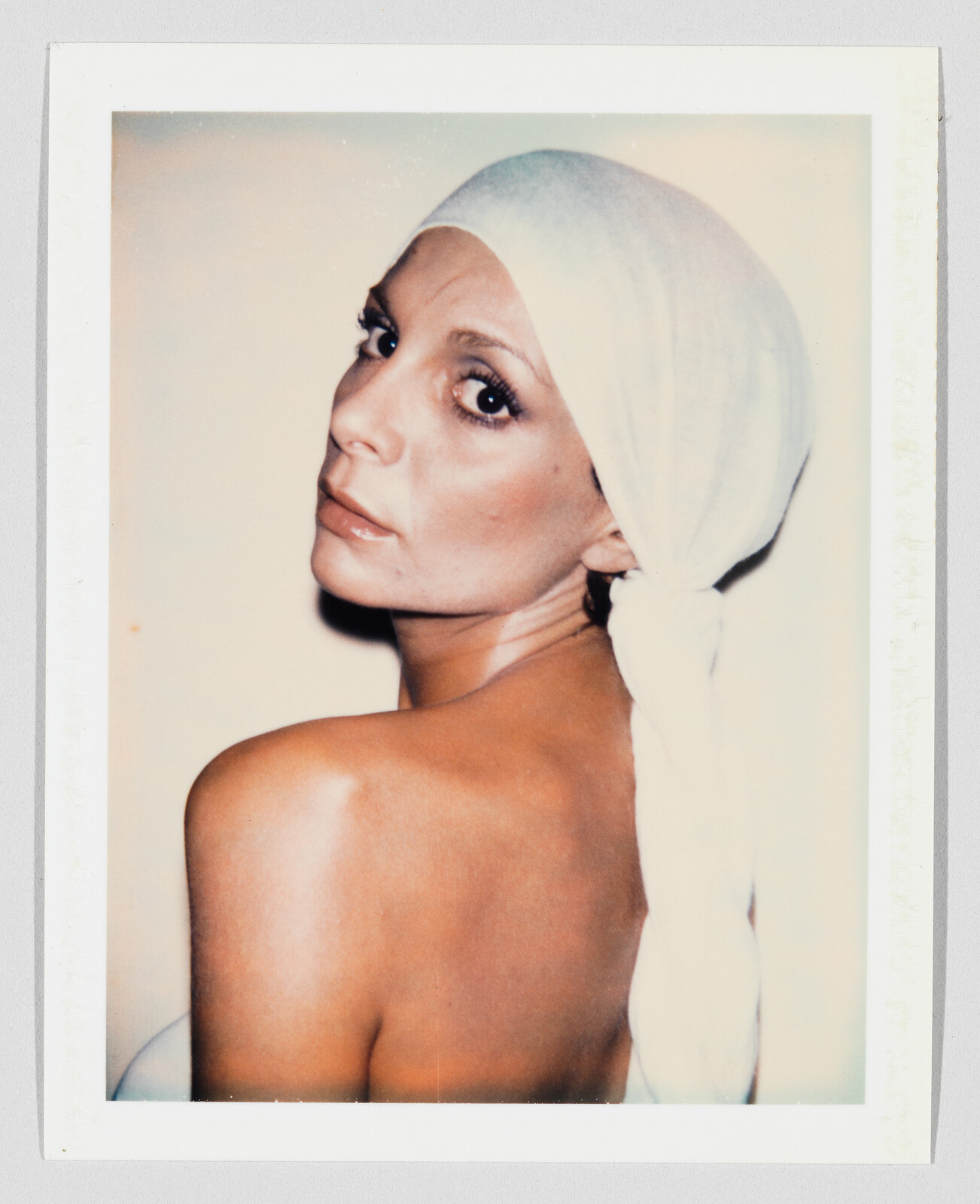 A woman with a white headscarf looks over her bare shoulder toward the camera.