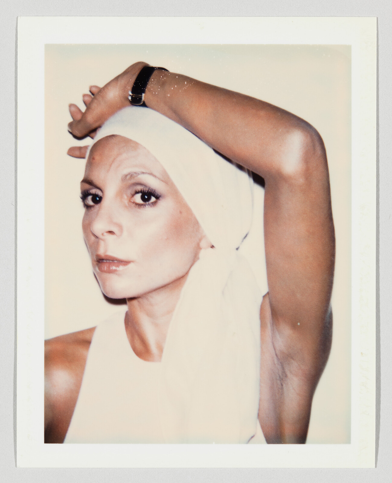 A woman in a white headscarf raises her arm, exposing her armpit while looking at the camera.