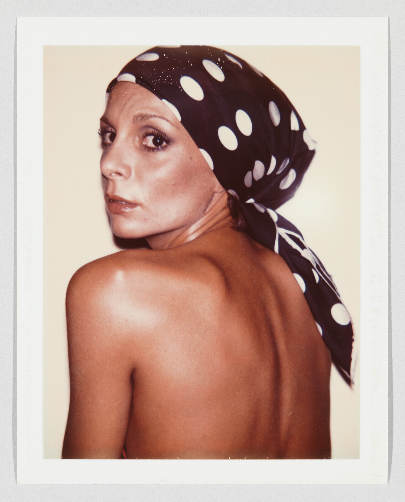 A woman with a polka dot headscarf looks over her shoulder, showing her bare back.