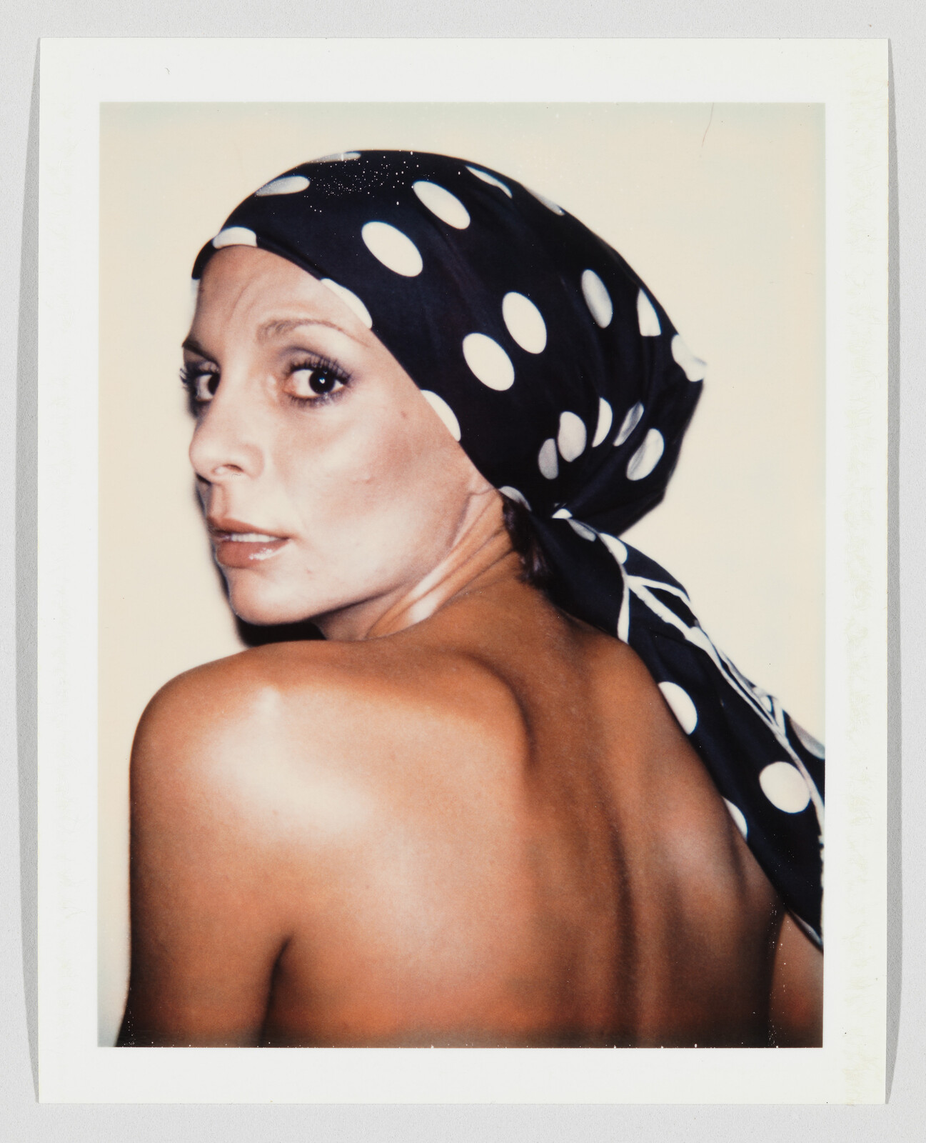 Woman with a black-and-white polka dot headscarf looks over her bare shoulder displaying her back.