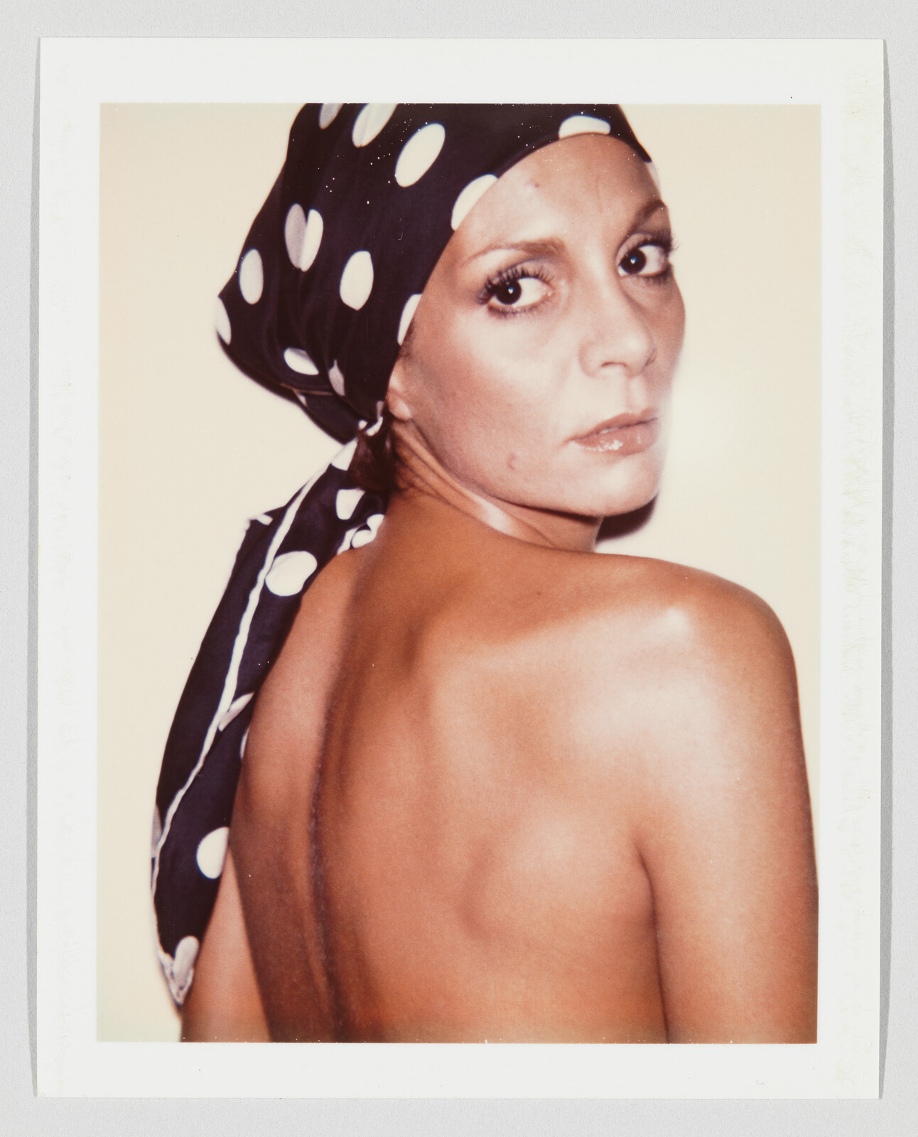 Woman wearing a black polka-dot headscarf looks over her shoulder showing a bare back.