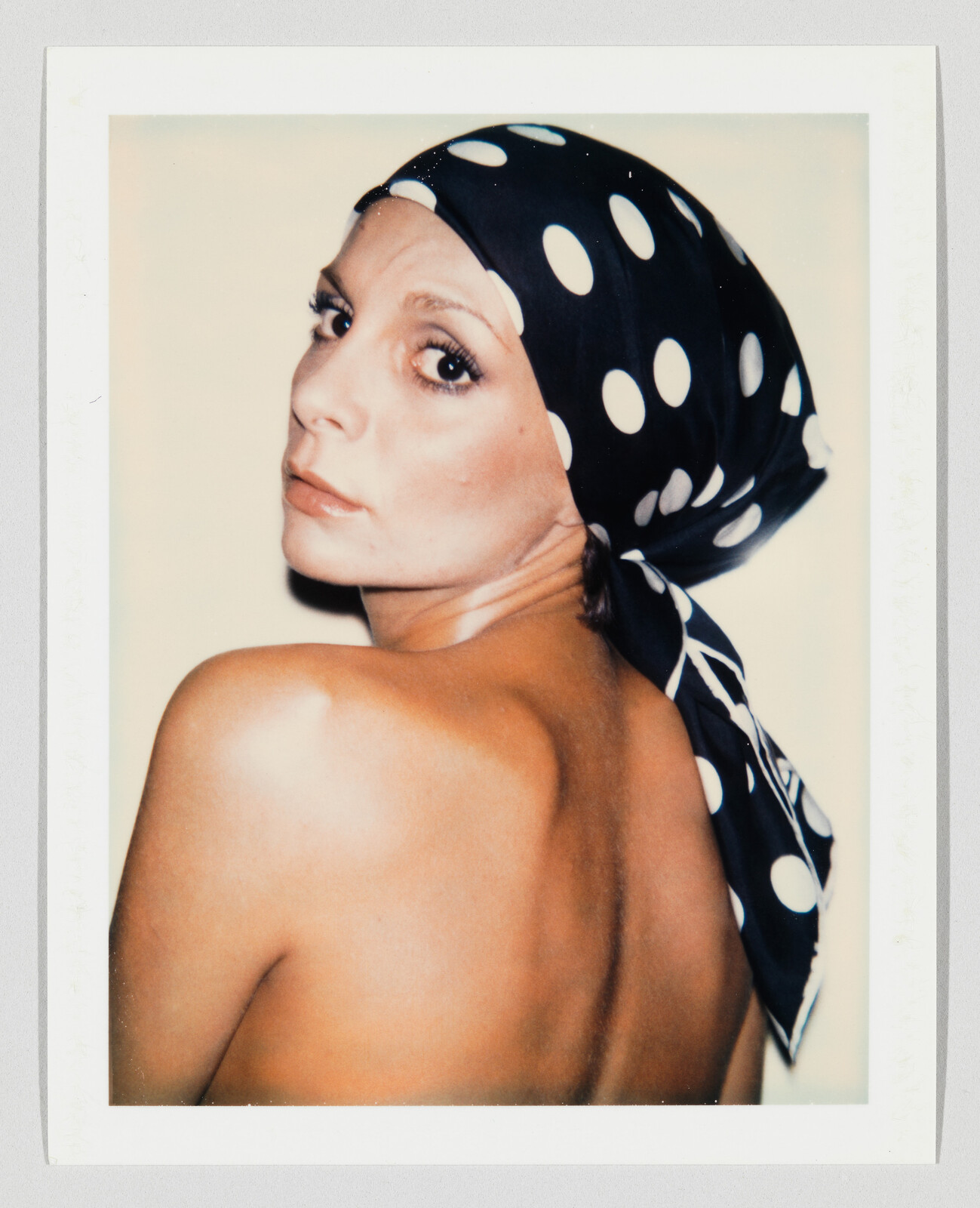Woman with bare shoulders looks over her shoulder while wearing a black polka-dot headscarf.