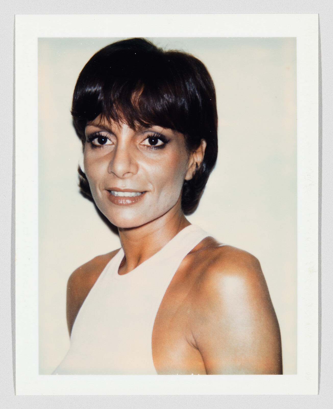 A woman with short dark hair smiles at the camera wearing a white sleeveless top.