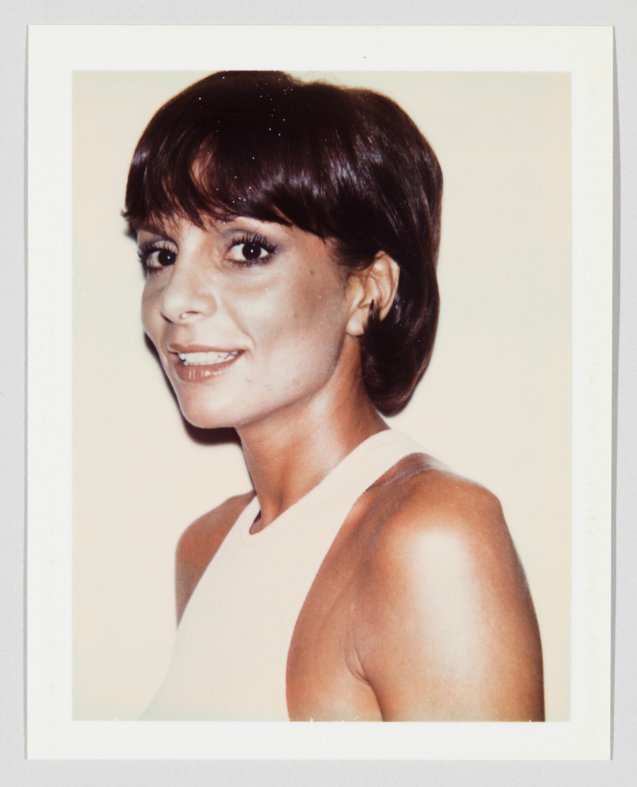 Woman with short dark hair smiling in a sleeveless white top, looking over her shoulder.