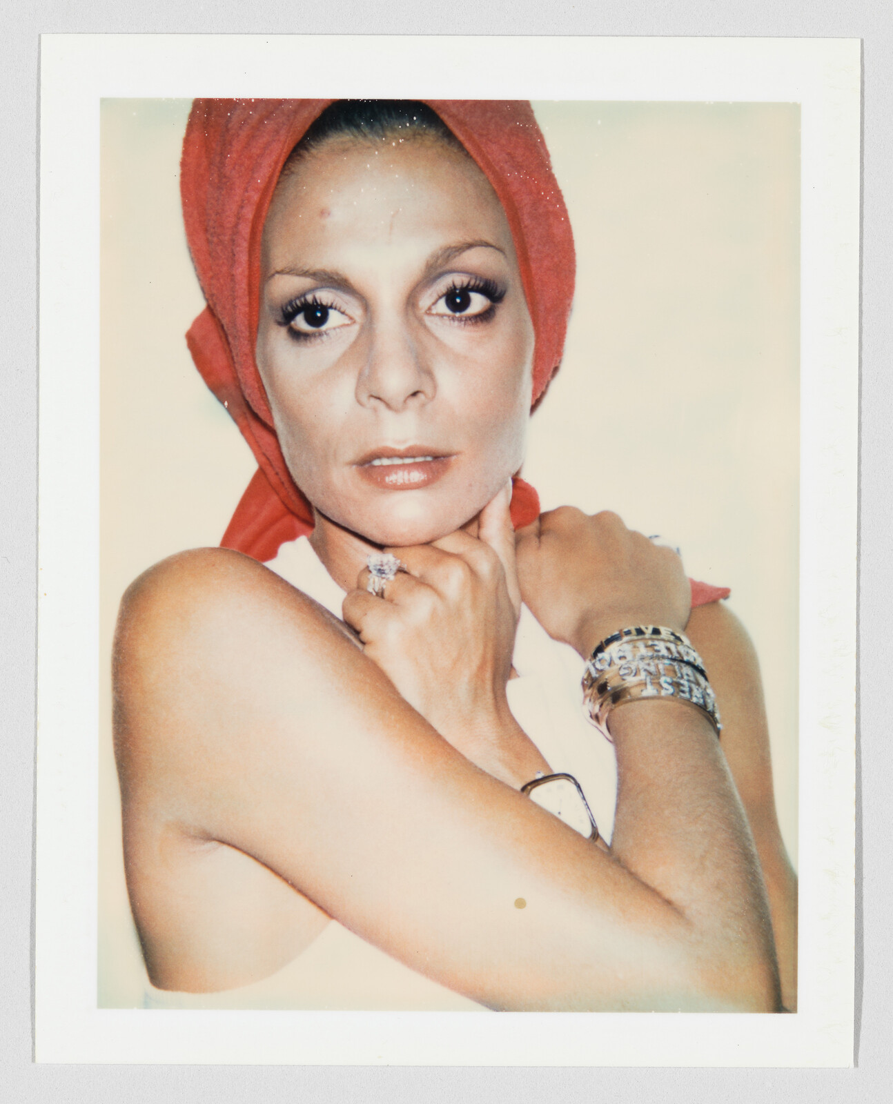 Woman wearing a red headscarf looks directly at the camera with arms crossed, showing jewelry.