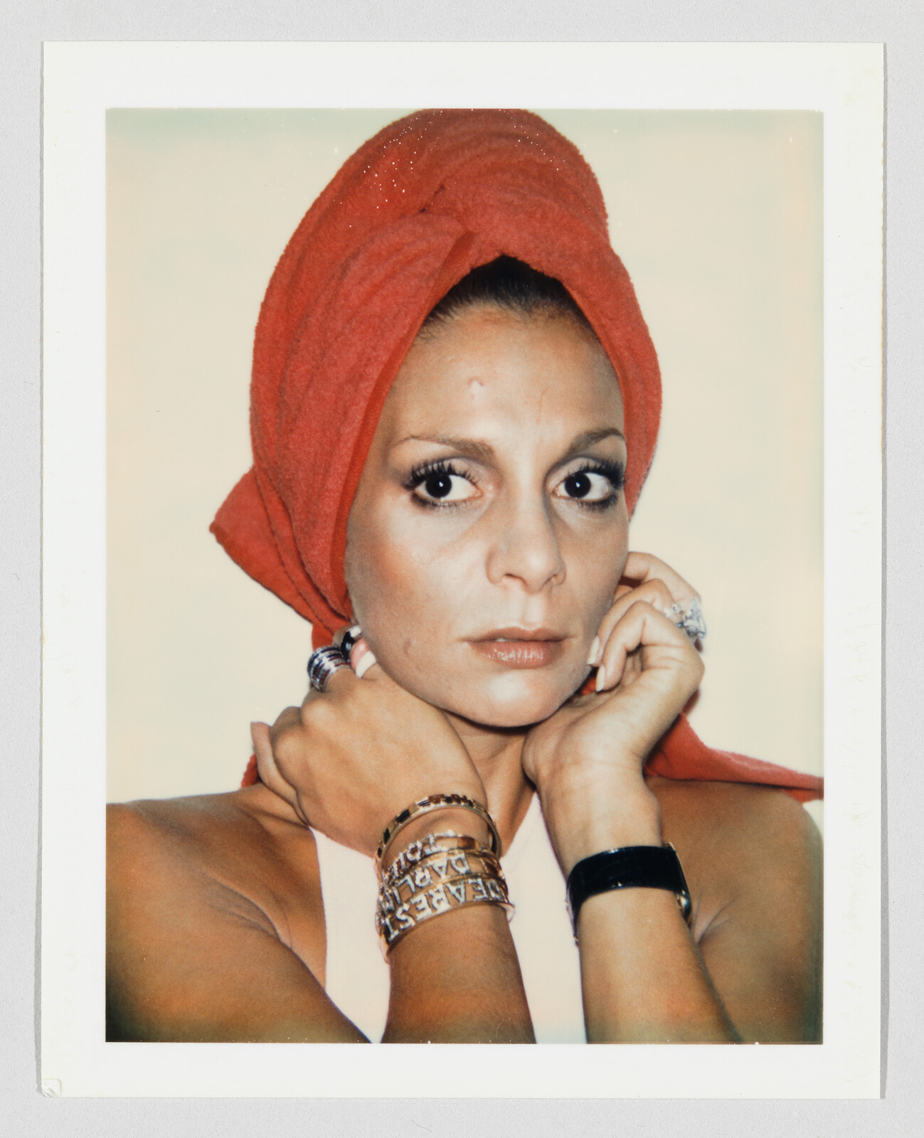 Woman wearing a red towel-wrapped turban looks straight at the camera while holding her neck, showing bracelets.