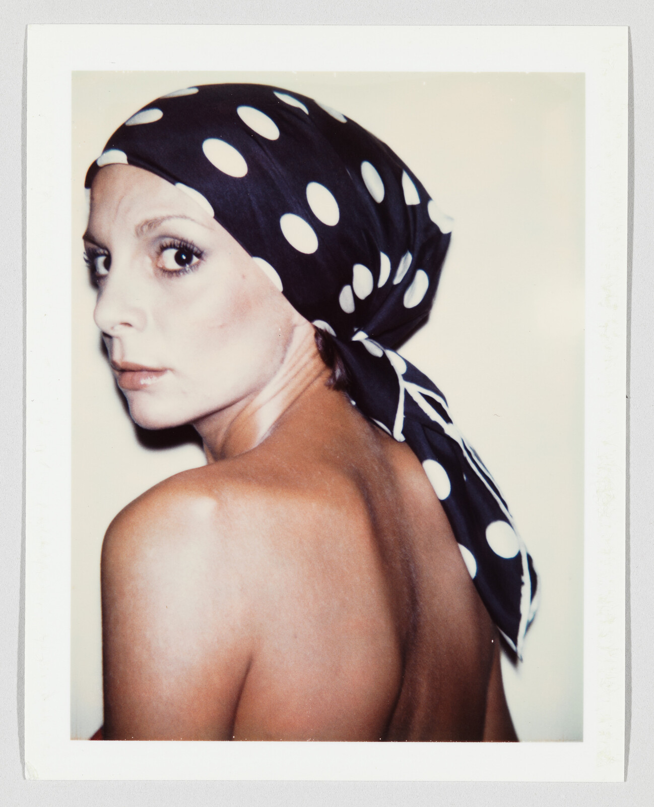 Woman wearing a polka-dot headscarf looks over her shoulder showing her bare back.