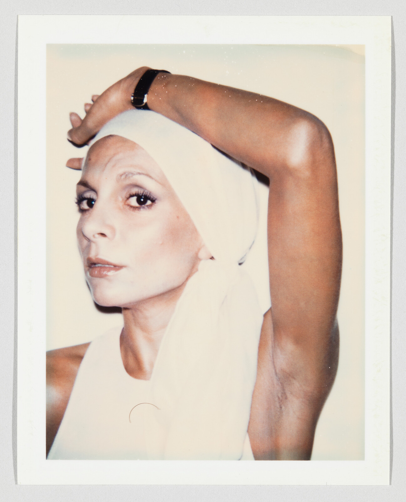 A woman wearing a white headscarf looks at the camera with one arm raised over her head.