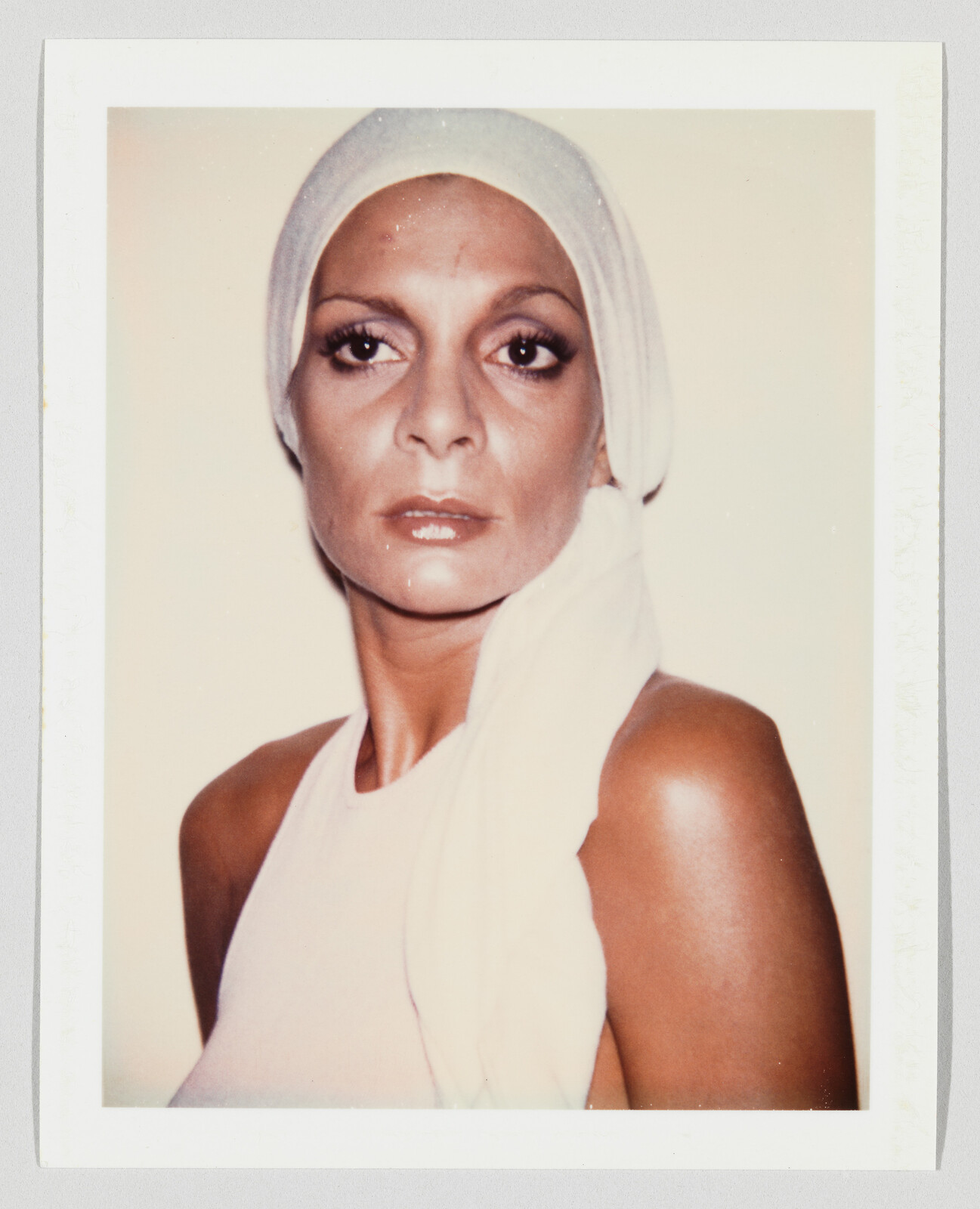 A woman wearing a white headscarf and sleeveless top looks straight ahead with a neutral expression.