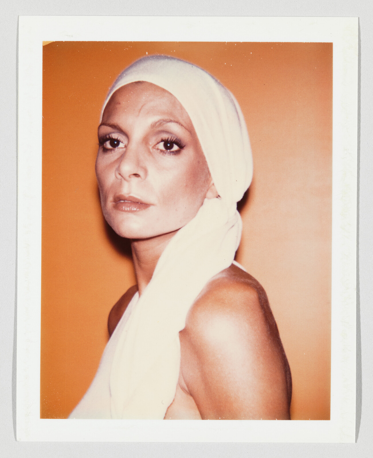 A woman with a white headscarf looks directly at the camera against an orange background.