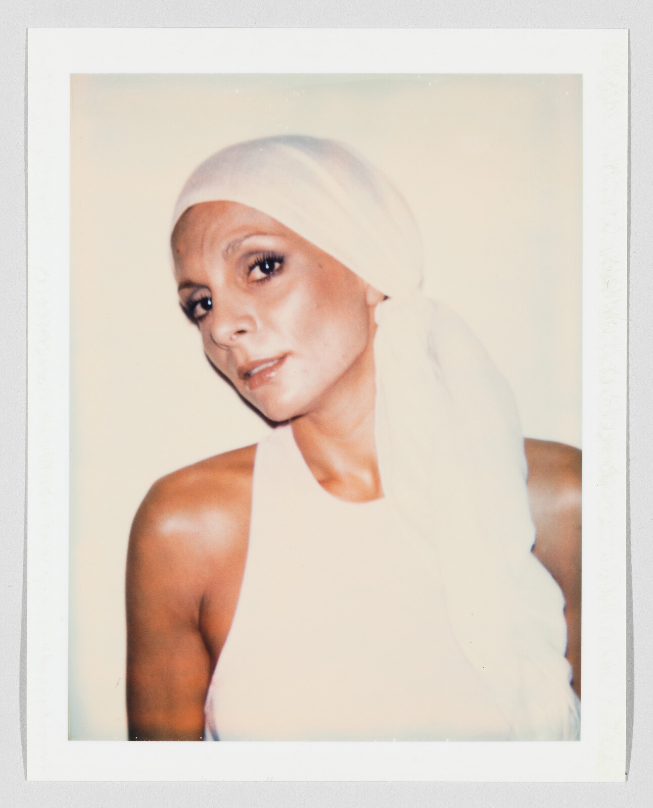 Woman wearing a white headscarf and sleeveless top looks softly toward the camera.