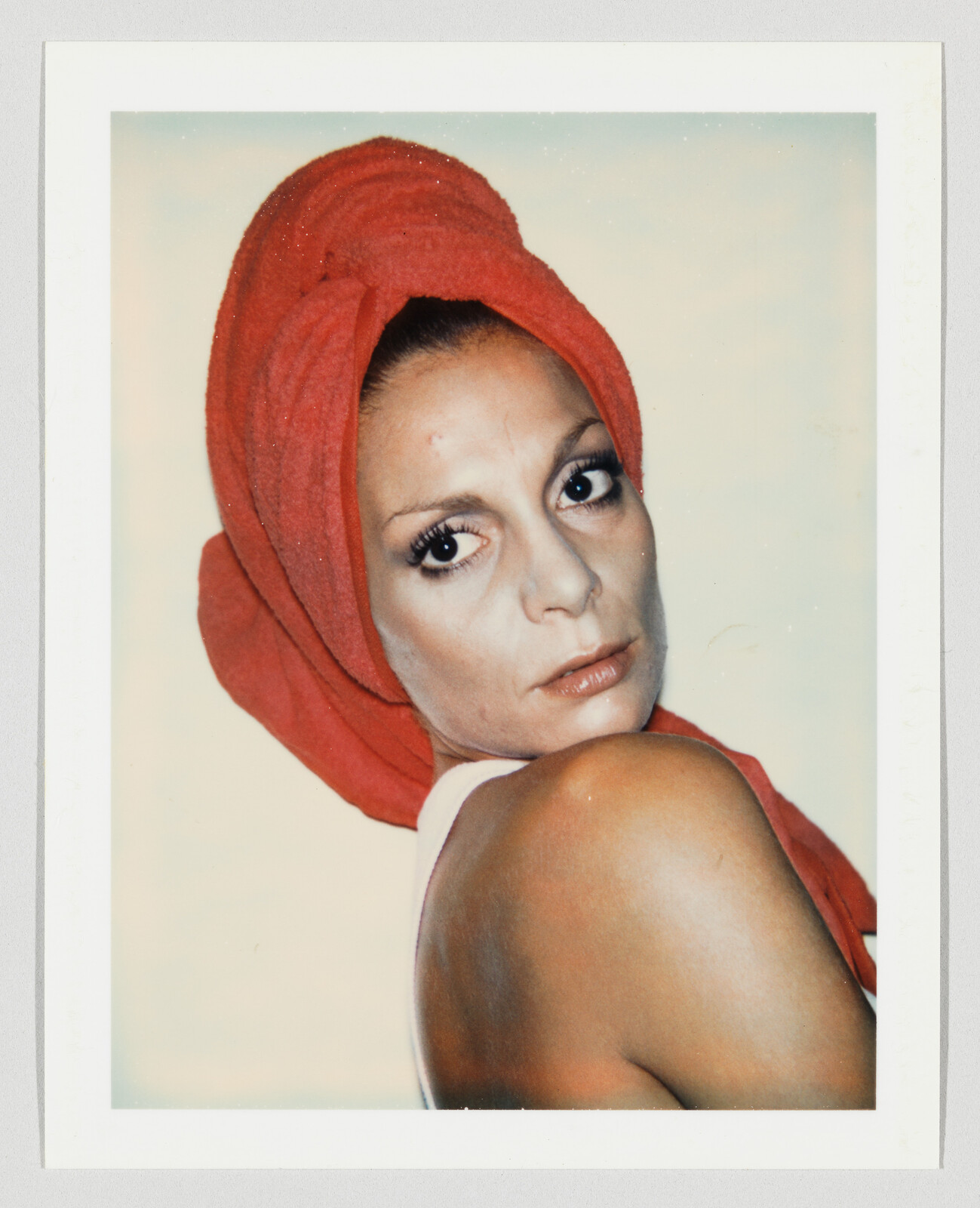 Woman with a red towel wrapped around her head looks over her bare shoulder at the camera.