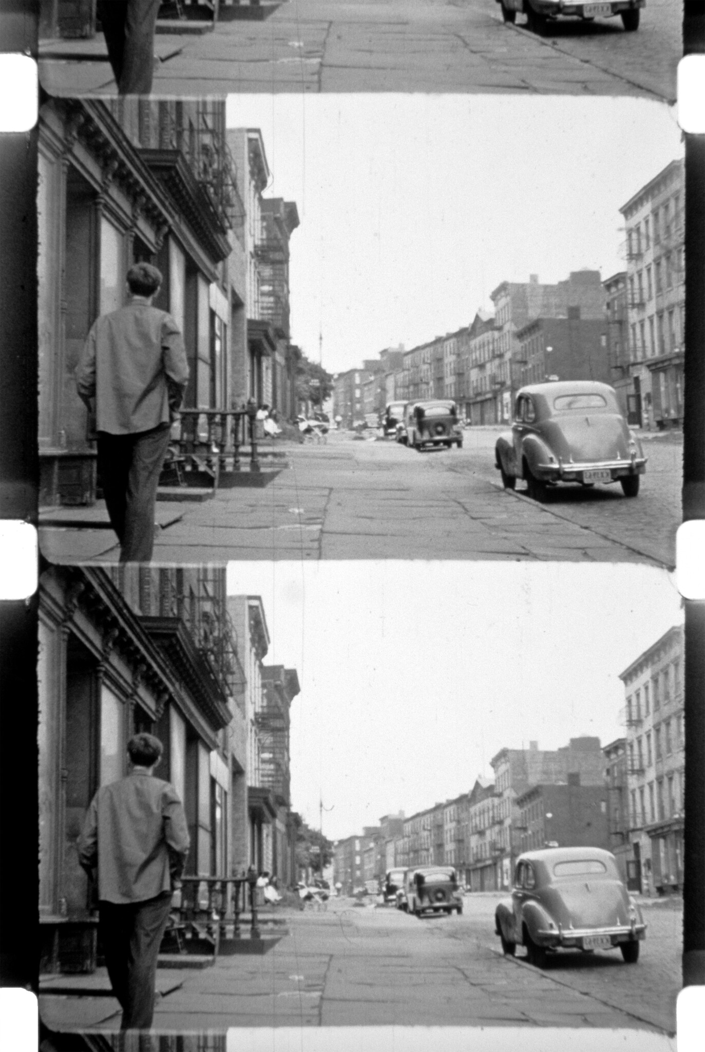 A man walks along a city sidewalk past parked vintage cars and brownstone buildings.