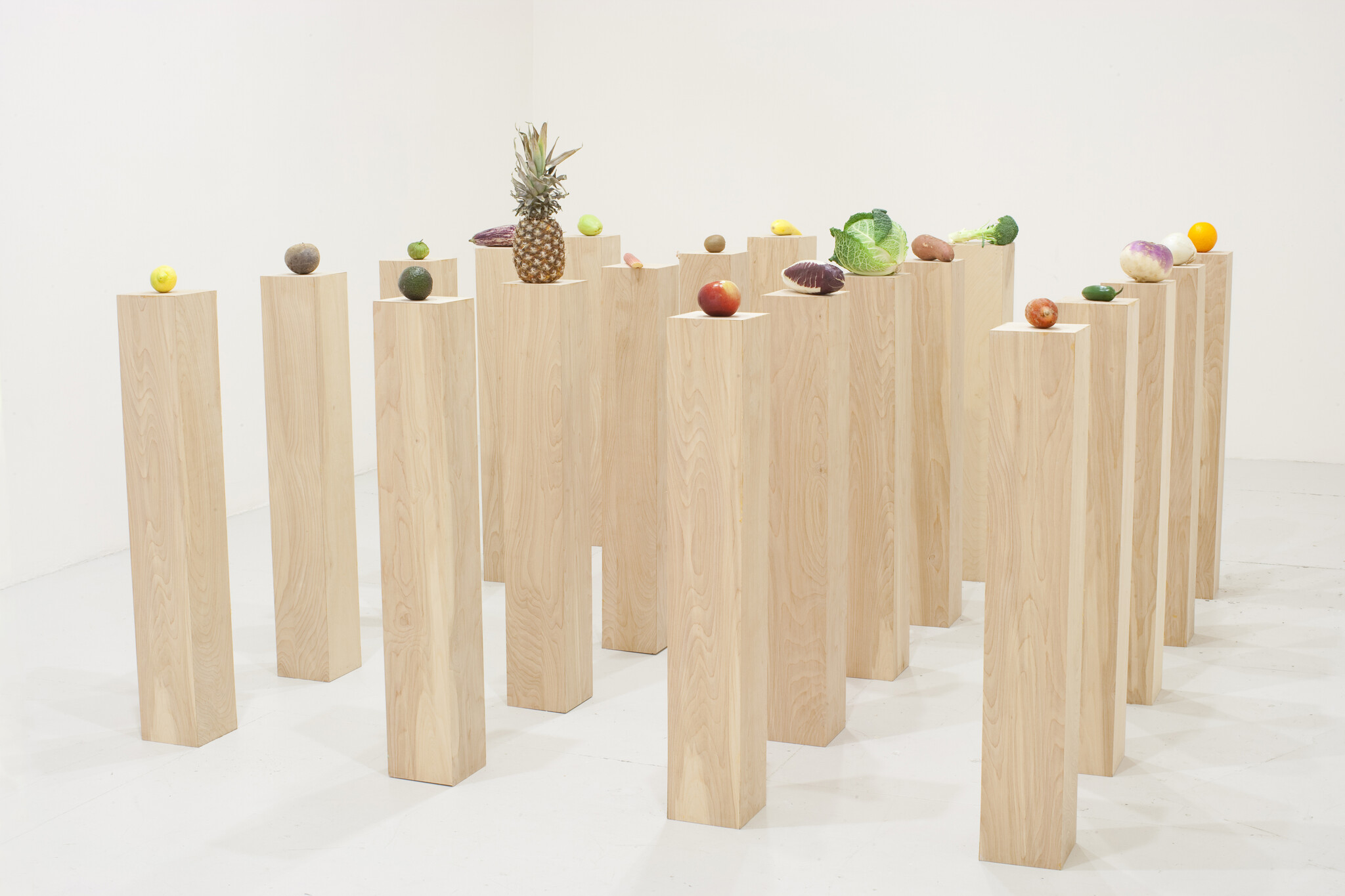 Multiple tall wooden pedestals arranged in rows each topped with a different fruit or vegetable.