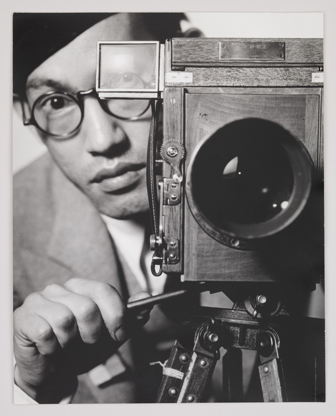 A person wearing glasses and a beret operates a vintage camera, focusing intently on the lens.