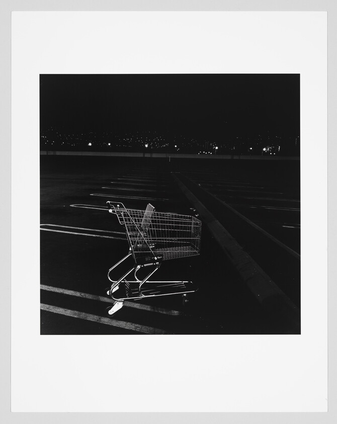 A lone shopping cart sits in an empty parking lot at night under distant lights.