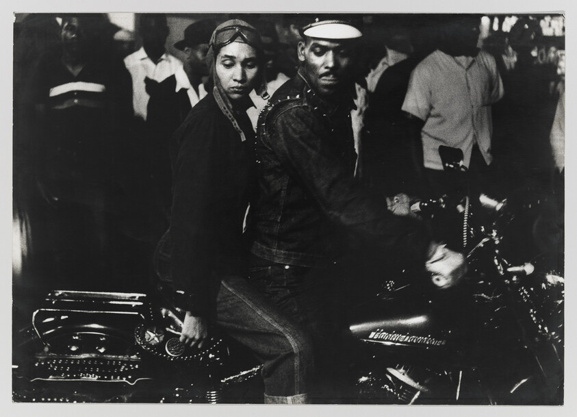 A man and woman sit on a parked motorcycle in a crowded street scene.