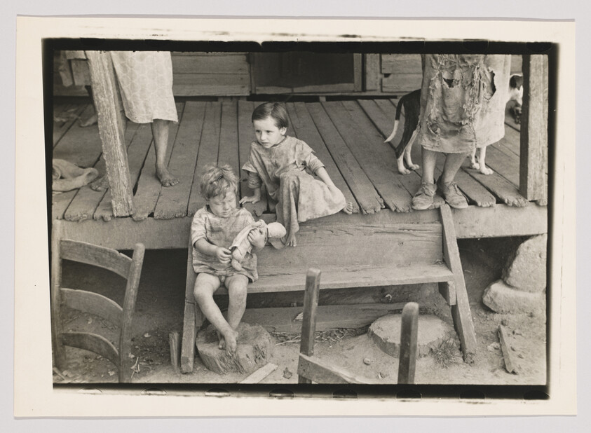 Two young children in worn clothes sit on wooden steps, with adults and a dog standing on the porch behind them.