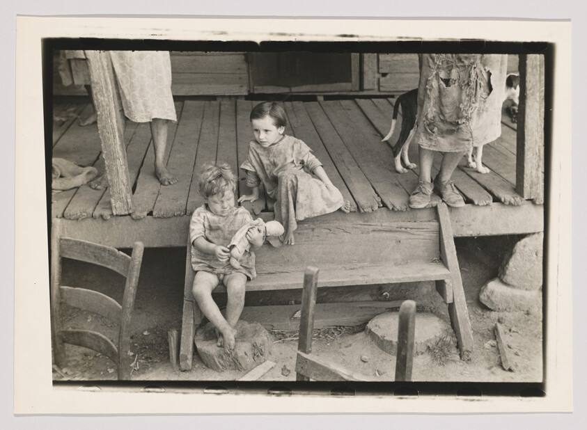 Two barefoot children sit on a worn wooden porch while one plays with a doll.