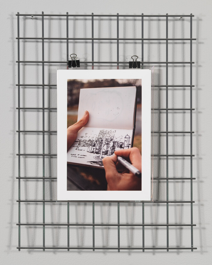 Two hands sketching a detailed pen drawing in a small sketchbook clipped to a wire grid.