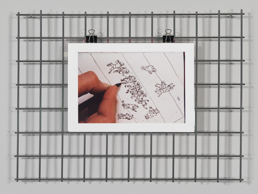 A hand draws tiny crowded human figures on a sheet of paper clipped to a metal grid.