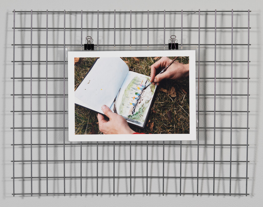 A photograph of a person's hands painting in a sketchbook is clipped to a metal grid on a wall. The painting depicts a colorful outdoor scene, and the sketchbook rests on a grassy surface. Shadows from the grid create a pattern on the wall behind.