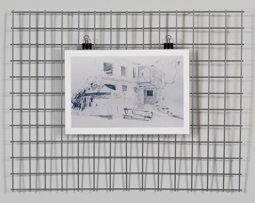 A sketch of a building scene is clipped to a metal grid on a white wall, casting a shadow behind it. The drawing is monochromatic and appears to be done in pencil, featuring the rough outline of a structure with windows and a sloped roof, possibly indicating an old house or barn.