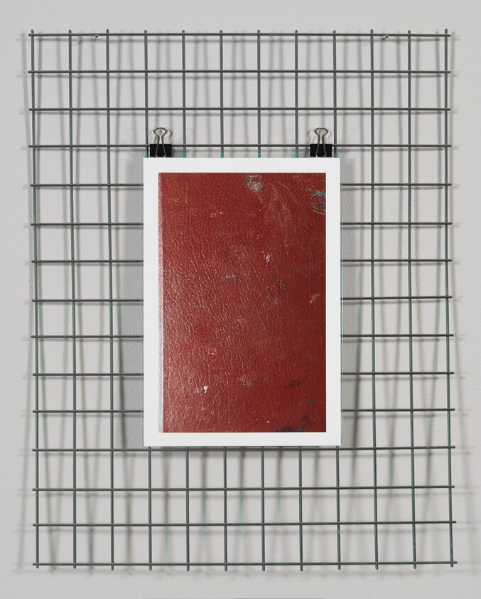A red textured artwork hanging on a metal grid against a white wall.