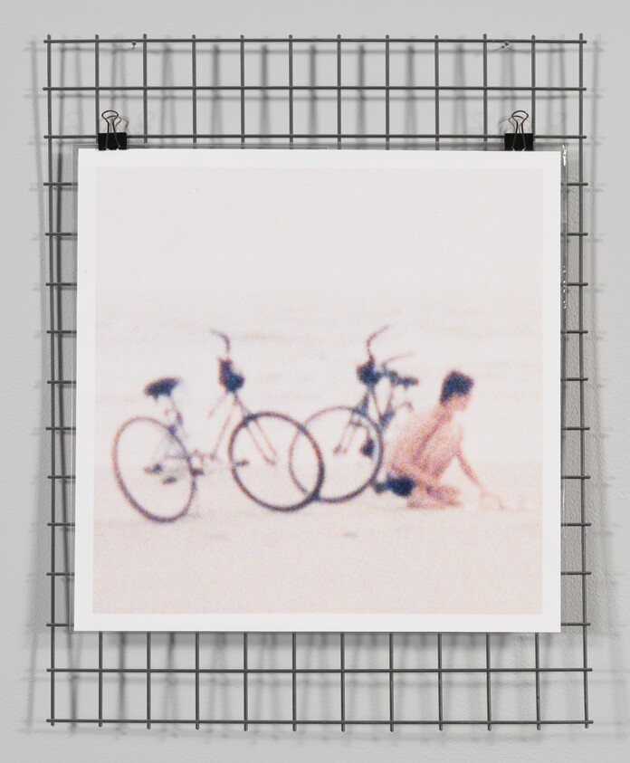 A blurred photograph of two bicycles and a person sitting on a beach, displayed on a white background and hung on a metal grid wall with clips.