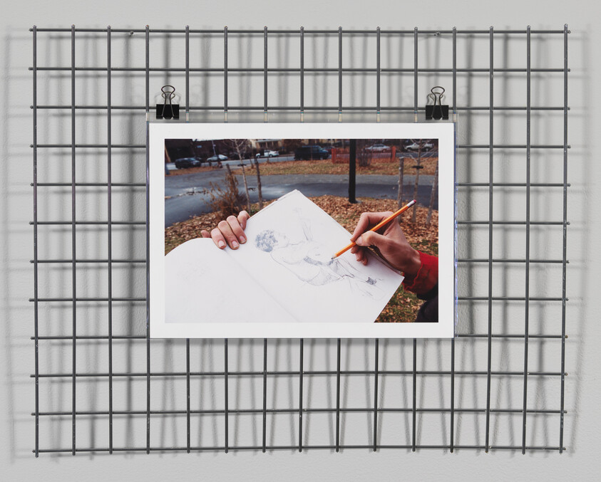 A photo of a person's hand drawing on a sketchpad is clipped to a metal grid on a wall. The drawing appears to be of a human figure, and the person is holding a pencil. The background of the photo within the photo shows an outdoor scene with bare trees and a park bench, suggesting the artist may be drawing from life.