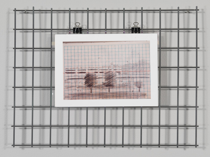 A piece of artwork with grid lines is clipped to a metal grid wall display using two black binder clips. The artwork features abstract smudges and lines, possibly in pencil or charcoal, on a background with a grid pattern that resembles graph paper. The metal grid is mounted on a light-colored wall, casting a shadow behind it.