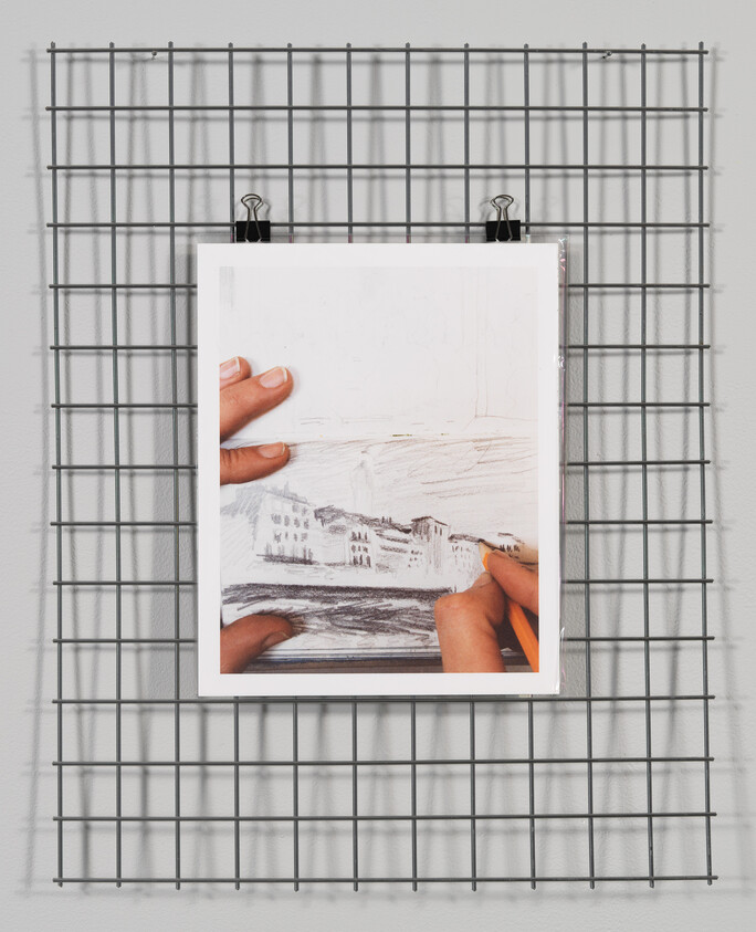 A sketch of a coastal scene with buildings is clipped to a wire grid on a wall. A person's hands are visible, one holding the sketch and the other using a pencil to draw. Shadows of the grid are cast on the wall.