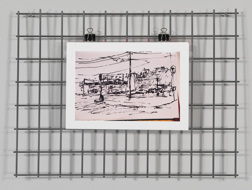 A black and white sketch of a street scene with utility poles and buildings is clipped to a metal grid on a white wall. The sketch has a rough, expressive style and is centered on a light pink paper with a red border on the bottom edge. Two black binder clips hold the artwork at the top. Shadows of the grid are cast on the wall, creating a geometric pattern.