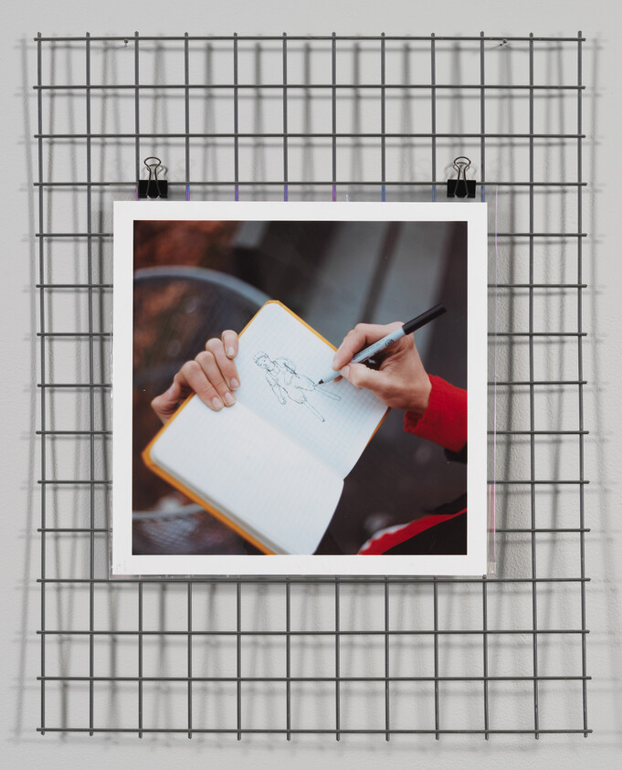 A photo of a person's hands drawing a figure on a notebook is displayed on a wire grid wall with clips.