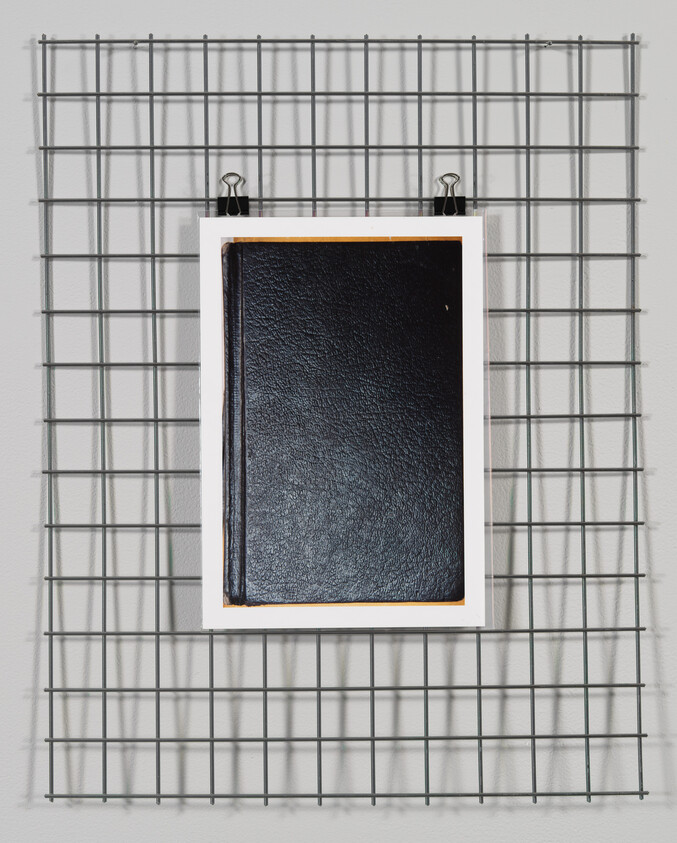 A closed black book with a white spine, clipped to a metal grid on a wall.