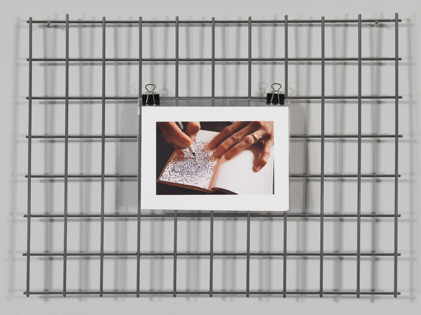 Photo of hands drawing on a notebook, clipped to a metal grid on a white wall.
