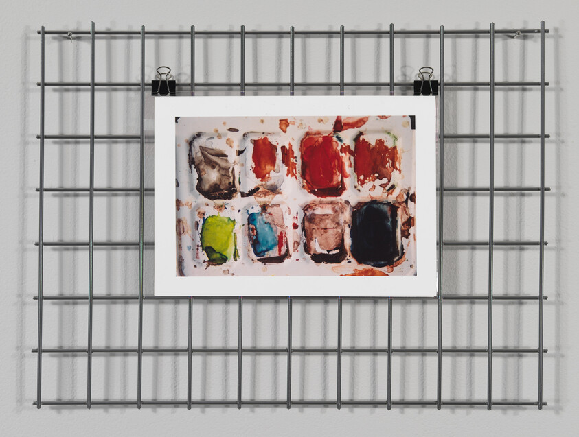 A watercolor paint palette with various colors hung on a metal grid wall by clips.