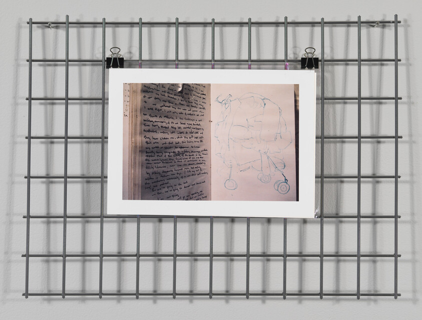 Artwork with handwritten text and abstract drawing clipped to a metal grid on a white wall.