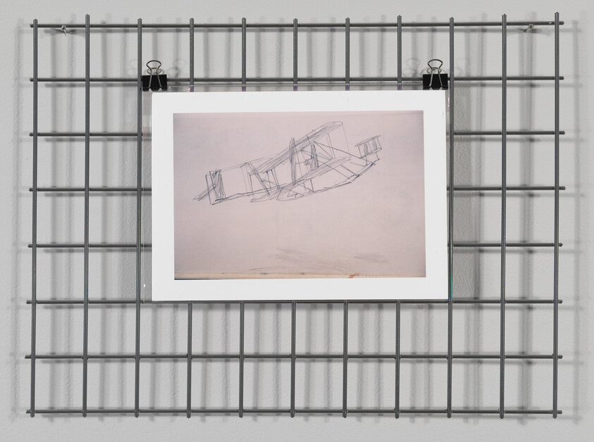 Sketch of an airplane on paper, hung on a metal grid wall with clips.