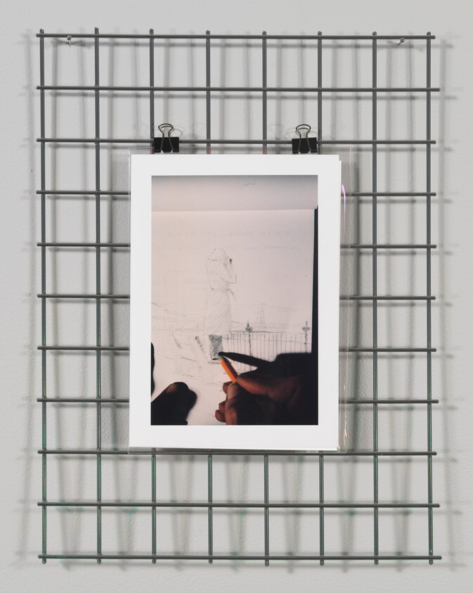 A sketch hangs on a metal grid wall using black binder clips. The drawing depicts a figure holding a pencil, appearing to sketch a scene with buildings. The artist's hand is visible at the bottom, suggesting a reflective surface on which the drawing is placed. Shadows of the grid cast onto the wall add depth to the image.