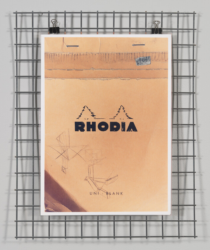 A worn Rhodia notepad hangs from two black binder clips on a metal grid wall. The notepad has visible creases and a blue stamp near the top edge. Below the brand name "RHODIA," there are abstract geometric shapes and the words "UNI_BLANK."