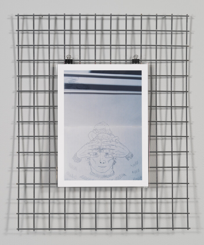 Sketch of a figure with hands on head hung on a metal grid against a white wall.