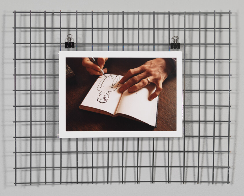 A person drawing a simple human figure in a small open notebook while holding the page.