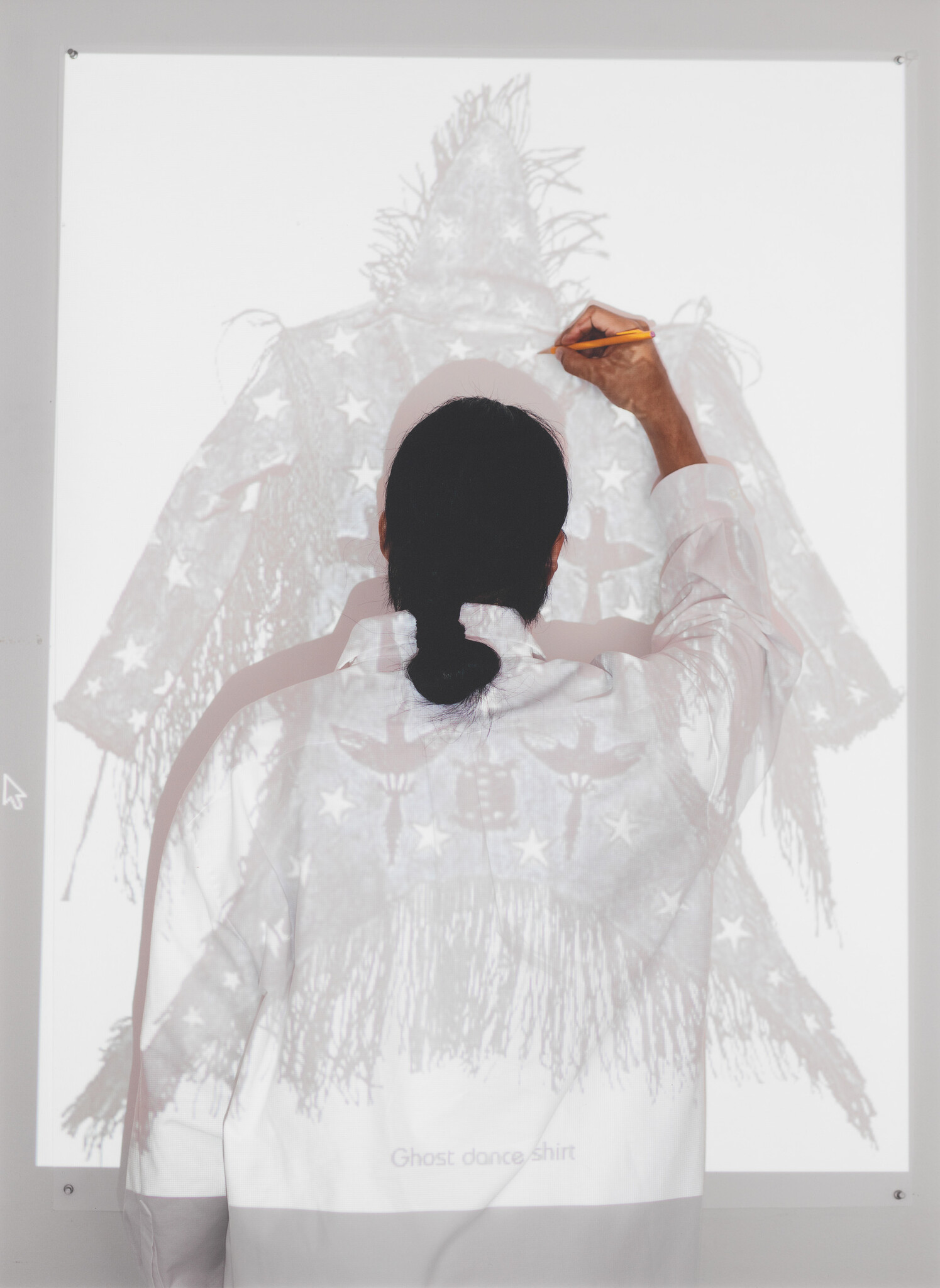 Person seen from behind tracing a projected ghost-dance shirt design with a pencil.