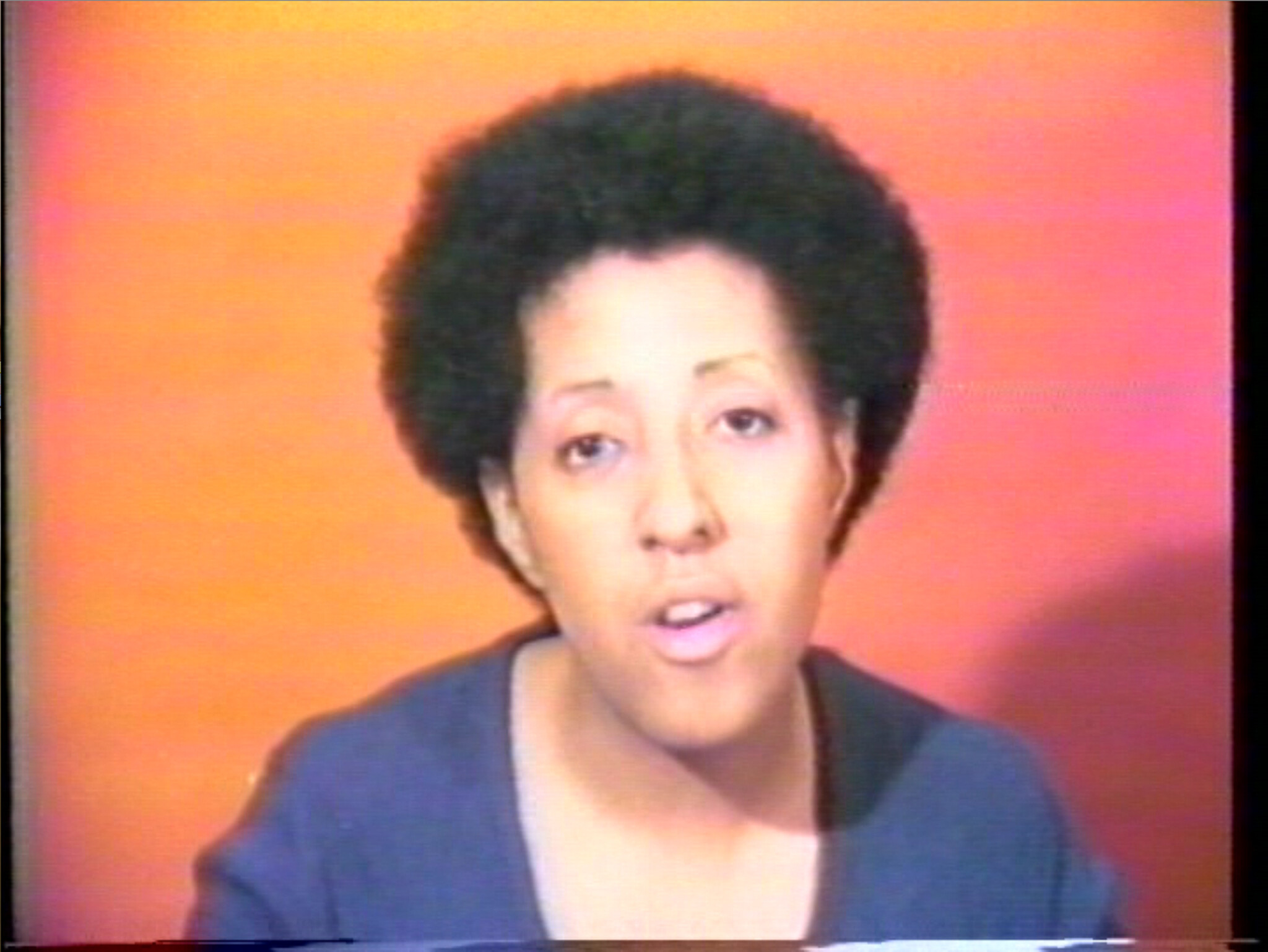 A person with a short afro hairstyle is in front of a red-orange background, appearing to speak or present. They are wearing a dark blazer over a lighter shirt. The image has a grainy quality, suggesting it may be from an older video source.