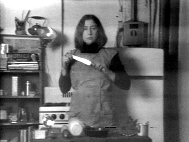 A woman in an apron holds a large kitchen knife while standing in a cluttered kitchen.